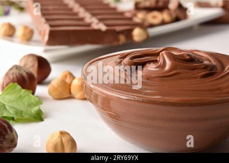 Primo piano della crema di cioccolato con nocciole in una ciotola su tavolo bianco con noci, cioccolato e foglie a vista. Vista anteriore rialzata. Foto Stock