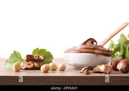 Crema al cioccolato con nocciole in ciotola e porzioni su tavola di legno con foglie e nocciole intorno e fondo bianco isolato. Vista frontale. Foto Stock