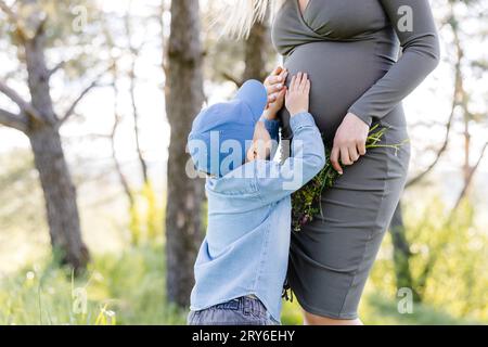 Ragazzo carino che accarezza il ventre incinta della mamma nella foresta. Madre incinta e suo figlio piccolo camminando e divertente trascorrere il tempo insieme Foto Stock