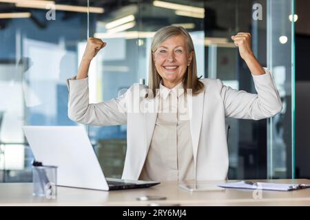 Ritratto di una donna d'affari anziana felice che lavora in ufficio utilizzando un notebook e si rallegra per il successo e i risultati ottenuti, sorride alla fotocamera e alza le mani per festeggiare. Foto Stock