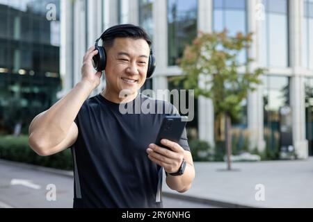 Giovane asiatico in uniforme sportiva in piedi in mezzo alla strada, indossa cuffie e regge un telefono, ascolta musica, audiolibro, podcast, fa sport. Foto Stock