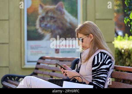 Donna con occhiali e smartphone seduta su una panchina in una strada cittadina su uno striscione con sfondo gatto Foto Stock