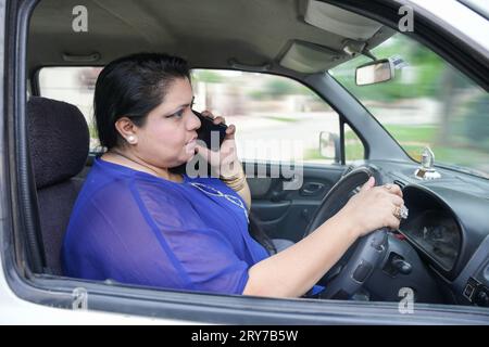 Giovane donna indiana di taglia più grande che parla al cellulare mentre si guida l'auto senza indossare la cintura di sicurezza, concetto di sicurezza. Foto Stock