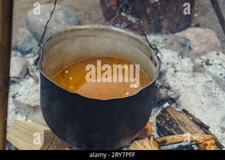 Recipiente di metallo con zuppa piccante e appetitosa appesa al treppiede sotto la legna da ardere prima di cucinare sul fuoco in natura. Pasto liquido all'arancia per prepa da campeggio Foto Stock