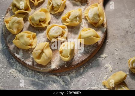 Tortellini ravioli tradizionali crudi su piatto di legno, tavolo grigio con farina Foto Stock