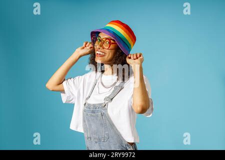 La donna sorridente con occhiali da vista indossa un berretto arcobaleno mentre la fotocamera si affaccia sullo sfondo blu dello studio Foto Stock