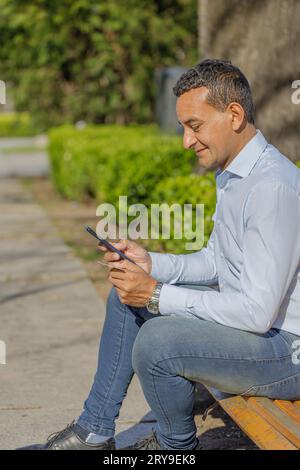 Un giovane latinoamericano che fa un acquisto con una carta di credito sul suo cellulare seduto su una panchina. Foto Stock