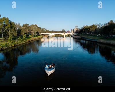 Vista di una piccola barca che corre lungo il fiume Yarra a Melbourne, nel tardo pomeriggio con alberi che incorniciano il fiume e un grande ponte nel Foto Stock