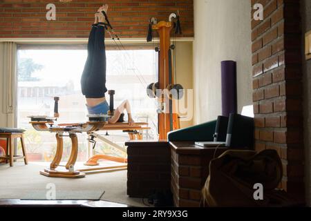 Donna caucasica con abbigliamento sportivo blu, nel soggiorno del suo appartamento, allenando la sua energia e l'equilibrio con la macchina per l'esercizio fisico, utilizzando pulegge con finestra Foto Stock