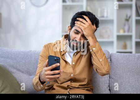 L'uomo ha ricevuto un messaggio di cattive notizie con notizie tristi online mentre era seduto sul divano in salotto utilizzando l'app sul telefono. Foto Stock
