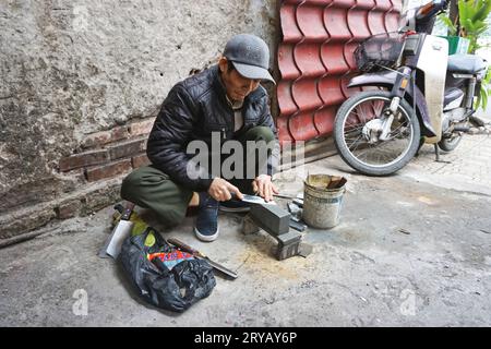 Un affilatore itinerante porta il suo mestiere al di fuori di un tradizionale mercato umido di Hanoi, in Vietnam. I venditori di carne all'interno sono i suoi clienti abituali. Foto Stock