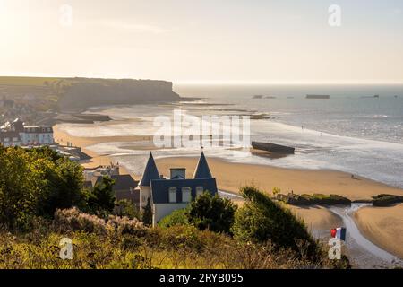 Vista sulla spiaggia di Arromanches in Normandia al tramonto con i resti del porto di gelso della seconda guerra mondiale che giace sulla sabbia e i cassoni Phoenix nel mare. Foto Stock