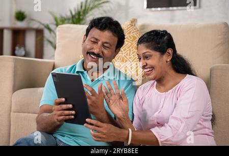 Felice e sorridente coppia indiana di mezza età che effettua videochiamate sul tablet digitale mentre si siede sul divano a casa: Concetto di connessione familiare, insieme Foto Stock