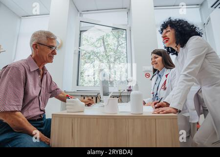 Un team sanitario diversificato che parla con un paziente maturo in un'atmosfera positiva, conduce test e analizza i risultati di laboratorio in una clinica. Concentrati su succ Foto Stock