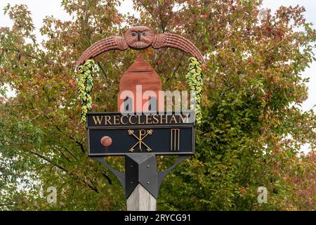 Cartello del villaggio di Wrecclesham, Surrey, Inghilterra, Regno Unito. Il segno decorativo presenta un forno per ceramiche, gufo e simboli di cricket che riconoscono le sue associazioni storiche Foto Stock