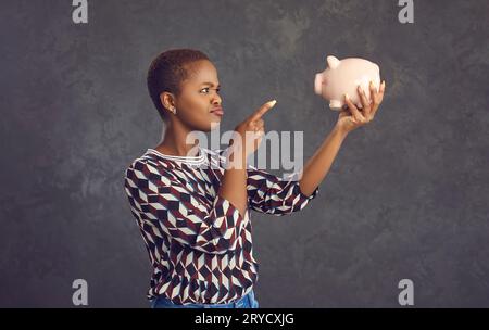 Una donna dalla pelle scura e spaventosa lancia un dito punitivo contro il salvadanaio facendo un gesto di vergogna. Foto Stock