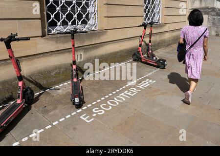 Una donna che cammina davanti a una baia di scooter elettrici in Beaumont Street, una prova del governo britannico di noleggio di scooter elettronici a emissioni zero nel centro di Oxford, in Inghilterra Foto Stock