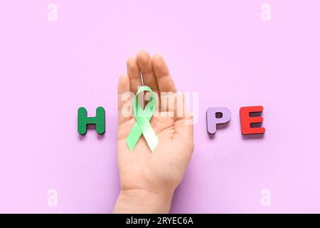 Mano femminile con nastro verde e parola HOPE fatta di lettere di legno su sfondo viola. Giornata della salute mentale Foto Stock