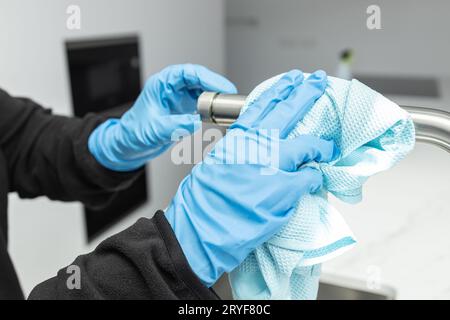 Mani in guanti protettivi pulizia di un rubinetto moderno con straccio di una cucina domestica minimalista Foto Stock