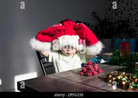 Un bambino piccolo con cappello di Babbo Natale e guanti rossi con regali di Natale è seduto al tavolo di Capodanno. Festeggiamo il Natale. Foto Stock