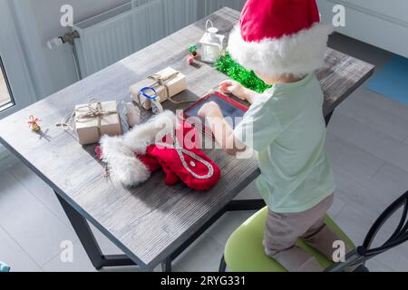 Un bambino piccolo con un cappello di Babbo Natale, con regali di Natale, è seduto a un tavolo di Capodanno con un tablet in mano. Festeggiamo il Natale. Foto Stock