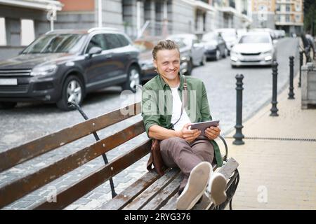 Uomo libero professionista felice che lavora utilizzando un tablet digitale siede sulla panchina all'aperto nell'edificio di front Office e indossa una camicia verde informale Foto Stock