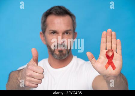 Pollice su uomo maturo con nastro rosso Bow AIDS simbolo di consapevolezza su un palmo che indossa una maglietta bianca isolata su uno schienale blu Foto Stock