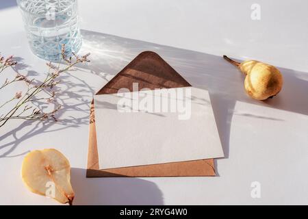 Busta con cartolina vuota, bicchiere d'acqua, pere e fiori alla luce del sole su un tavolo bianco. Vista dall'alto, base piatta, mockup. Foto Stock