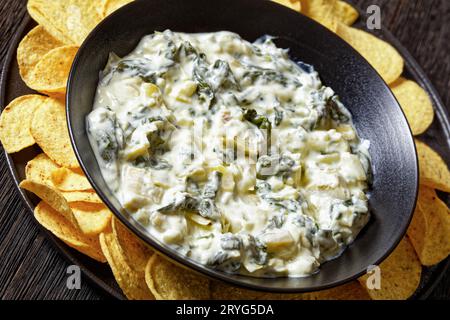 primo piano della salsa cremosa di carciofi spinaci alle mandorle con patatine fritte in ciotola su piatto nero su tavolo di legno scuro, angolo olandese Foto Stock