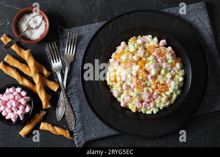 Insalata Ambrosia con ananas, arance mandarine, yogurt, mini marshmallow, cocco e panna montata in una ciotola nera su un tavolo di cemento con ingredienti Foto Stock