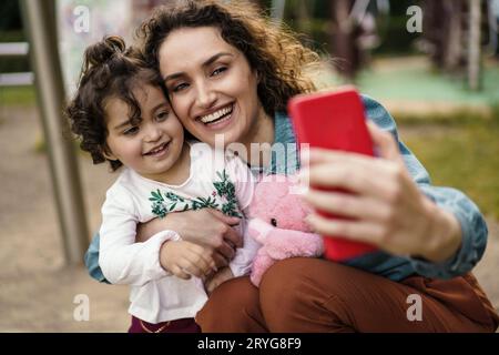 Donna brasiliana di 40 anni, con lunghi capelli ondulati, si accovaglia per abbracciare sua figlia per un selfie in un parco pubblico. Entrambi sorridono brillantemente, con la bambina Foto Stock