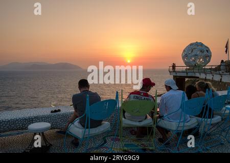 IOS, Grecia - 8 settembre 2023: I turisti si godono un drink e una vista mozzafiato sul Mar Egeo con un tramonto arancione in un lounge bar in Grecia Foto Stock