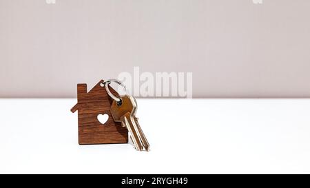 Chiavi metalliche per la casa in un portachiavi in legno a forma di casa con un foro a forma di cuore sopra un tavolo avvolgente di fronte a una parete grigio chiaro. Foto Stock