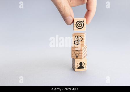 Strategia del team aziendale e concetto di piano d'azione. Mano che posiziona il blocco di legno con l'icona del bersaglio sopra gli altri con l'ingranaggio, lampadina Foto Stock