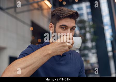 Primo piano di un bell'uomo barbuto che beve caffè su uno sfondo moderno di coworking e sembra una fotocamera Foto Stock