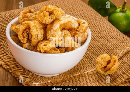 Vista dall'alto delle croste di maiale fritte e croccanti fatte in casa in una ciotola di ceramica bianca su un panno da sacco. Foto Stock