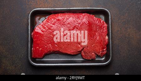 Bistecca di manzo rotonda cruda non cotta con sangue in vassoio di polistirene nero del supermercato su fondo rustico marrone scuro fr Foto Stock