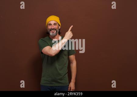 L'eccitato uomo caucasico sorridente con un cappello giallo punta la mano di lato con uno spazio per la copia su sfondo marrone. Posizionamento del prodotto Foto Stock