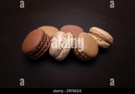 Macaron di colore beige, marrone e crema su sfondo di ardesia nera. Primo piano in diverse posizioni. Messa a fuoco selettiva. Foto Stock