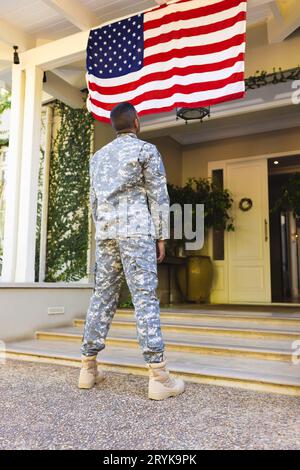 Biracial maschio soldato americano indossando uniforme militare guardando la bandiera degli stati uniti fuori dalla casa Foto Stock