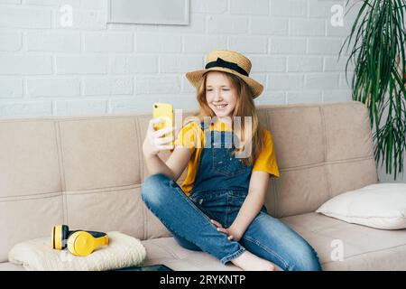 Allegra ragazza rossa in un cappello di paglia e tute in denim che posano e prendono un selfie su uno smartphone. Foto Stock