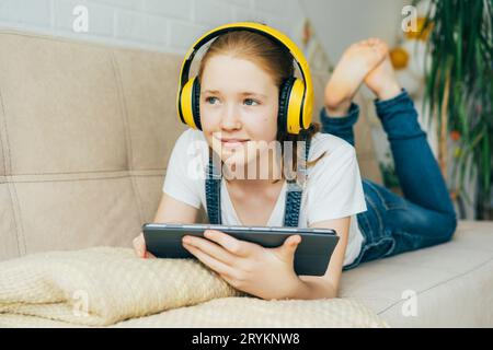 Una bella ragazza rossa sdraiata sul divano con un tablet elettronico ascolta la musica con le cuffie gialle. Apprendimento remoto Foto Stock