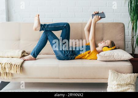 La ragazza adolescente in tuta in denim si rilassa sul divano con un tablet elettronico. Il bambino comunica in videochat Foto Stock