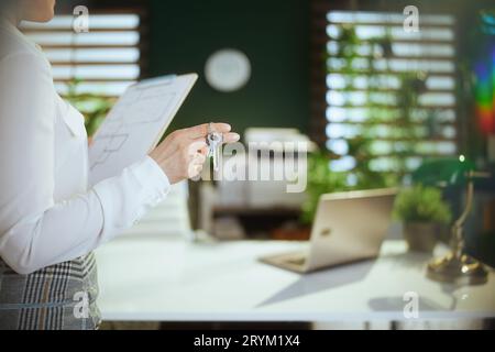 Un'attività immobiliare sostenibile. Primo piano su una giovane agente immobiliare in un moderno ufficio verde con blusa bianca con appunti e chiavi. Foto Stock