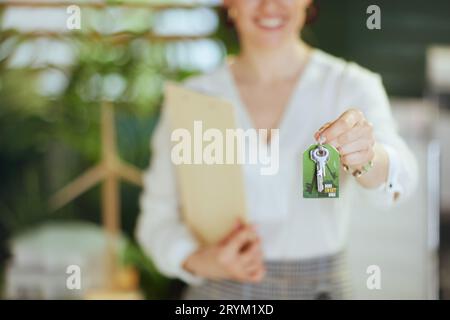 Un'attività immobiliare sostenibile. Primo piano su un moderno agente immobiliare femminile in un moderno ufficio verde con blusa bianca con clipboard e chiavi. Foto Stock