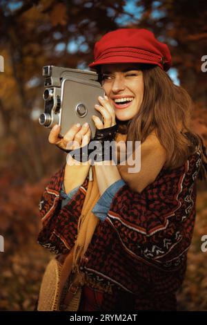 Ciao autunno. donna moderna sorridente di 40 anni con cappello rosso con videocamera retrò, sciarpa e guanti nel parco cittadino. Foto Stock