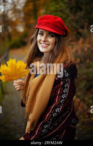 Ciao autunno. felice ed elegante donna di 40 anni in cappello rosso con foglie autunnali e sciarpa che cammina nel parco cittadino. Foto Stock