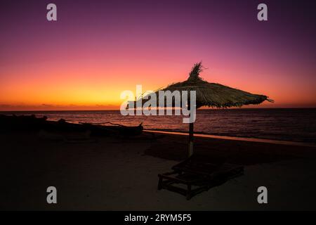 Vivace cielo rosso al tramonto sulla spiaggia di Anakao in Madagascar, con un ombrellone tropicale e un lettino Foto Stock