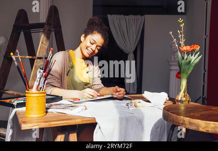 La giovane artista afro-americana, creativa e graziosa, dipinge con i colori pastello mentre si siede al tavolo davanti alla finestra Foto Stock