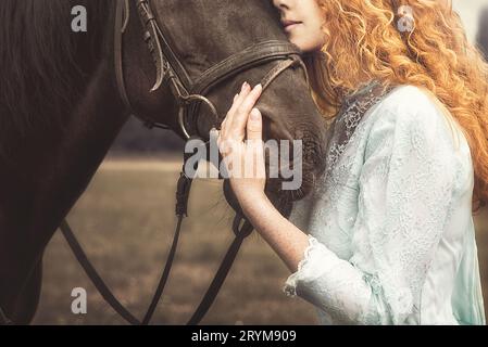 Foto artistica. Giovane donna senza volto vestita con un elegante abito vintage, coccola delicatamente un cavallo, accarezzandogli la testa. amore f Foto Stock